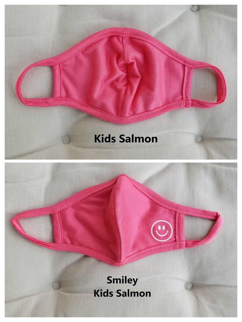 May include: Pink kids face mask with a white smiley face design. The mask is made of a soft, breathable fabric and has adjustable ear loops.