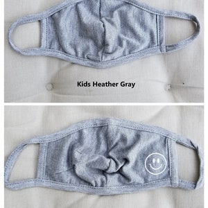 May include: Two gray face masks for kids. The top mask is plain, while the bottom mask has a white smiley face design. Both masks are labeled "Kids Heather Gray".