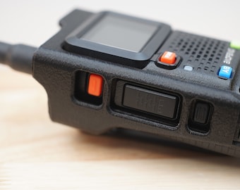 Pode incluir: Um rádio bidirecional preto com um botão laranja e um interruptor deslizante preto. O rádio tem uma tela pequena e um teclado.