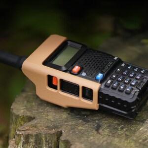 May include: A black and tan two-way radio with a tan protective case. The radio has a keypad, a screen, and a small antenna. The radio is sitting on a tree stump.