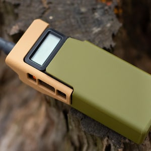 May include: A tan and olive green handheld device with a small screen and antenna. The device has a rectangular shape with rounded edges. The device is lying on a piece of wood.