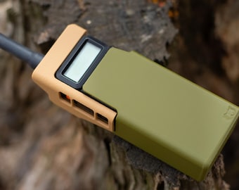 May include: A tan and olive green handheld device with a small screen and antenna. The device has a rectangular shape with rounded edges. The device is lying on a piece of wood.