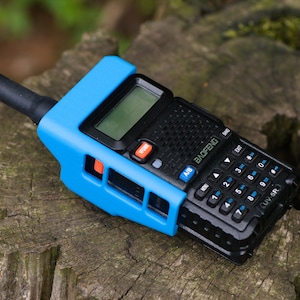 May include: A black and blue two-way radio with a blue protective case. The radio has a small screen and a keypad with numbers and symbols. The radio is labeled "BAOFENG" and "UV-5R".