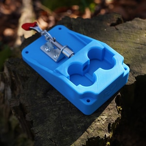 May include: A blue plastic 3D printed device with a red lever and two circular openings. The device is sitting on a tree stump.