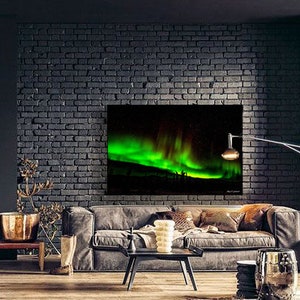 May include: A green and black canvas print of the aurora borealis, or northern lights, hanging on a brick wall in a living room. The print is above a gray couch with pillows and a coffee table.