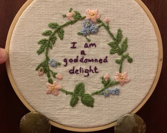 Könnte beinhalten: Stickrahmen-Kunst mit einer gestickten Blumenkrone und dem Text "I am a goddamned delight" in lilafarbenem Garn.
