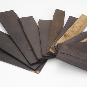 May include: A selection of dark brown wooden planks, of different sizes and shapes, arranged against a white backdrop. Some planks have lighter, tan-coloured sections, revealing the natural wood grain. The layout implies a display of wood samples or craft materials.