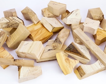 May include: A collection of assorted raw wood blocks and offcuts in various shapes and sizes. The pieces feature natural light tan and cream wood grains, some with dark brown bark edges, knots, and handwritten labels on their surfaces.