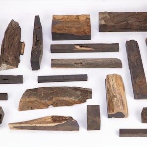 May include: A collection of various wooden blocks and irregular pieces of dark brown timber. The assortment includes rectangular bars, square blocks, and raw, rough-edged wood segments with visible grain patterns and natural textures in shades of brown.