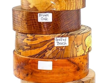 May include: A vertical stack of nine cylindrical wooden blocks with varying natural grain patterns and warm brown tones. Each block features a small white label with handwritten text identifying the wood type, including Mulberry, Spalted Silver Birch, Spalted Elm, Brown Oak, Spalted Beech, Holly, Burr Oak, and Spalted Holm Oak.