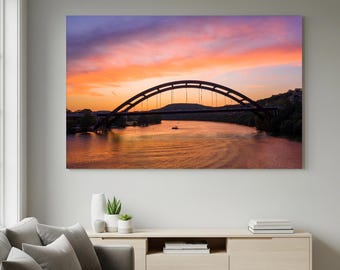 May include: A large canvas print of a bridge over a river at sunset. The bridge is dark brown and arched. The sky is a mix of orange, pink, and purple. The river reflects the colors of the sky. The print is hanging on a light gray wall.