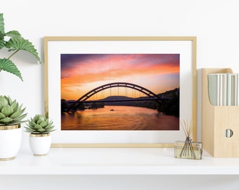 May include: A framed photograph of a bridge over a river at sunset. The sky is a vibrant mix of pink, orange, and purple. The bridge is silhouetted against the sky, and the water is a calm, reflective surface.