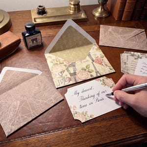 May include: A set of stationery featuring envelopes with a Paris-themed floral and Eiffel Tower print, alongside envelopes with a brown map pattern. A white card with floral accents displays the handwritten text, My dearest, Thinking of our time in Paris.