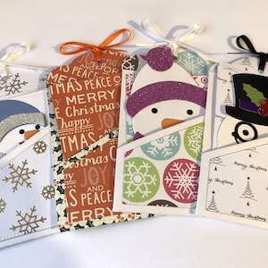 Puede incluir: Cuatro etiquetas de regalo navideñas hechas a mano con diseños de muñecos de nieve. Cada etiqueta tiene un bolsillo con un fondo estampado diferente y una cinta para colgar. Los sombreros de muñeco de nieve son azules, morados y negros.