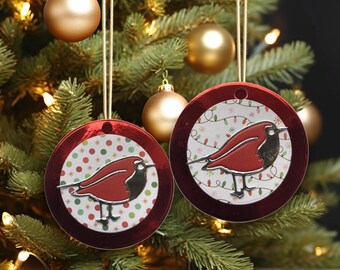 May include: Two circular Christmas tree decorations featuring a red robin design. The robin has a red body, black head, and silver details. The background is white with a pattern of lights and dots, framed by a red border. They hang from gold strings.