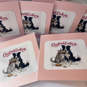 May include: A collection of square greeting cards with light pink borders. Each card features a central illustration of a gray cat and a black and white dog tangled in string lights, sharing a cookie. The text Christmas Cookies is printed above.