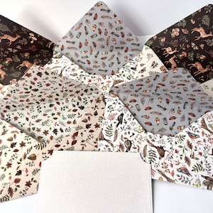 May include: Several open envelopes with nature-inspired patterns. The designs feature deer, leaves, feathers, and birds in brown, beige, and grey tones. A blank, white card is visible in the foreground.