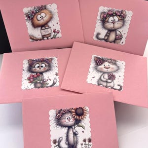 May include: Pink greeting cards featuring whimsical cat illustrations with floral crowns. Each card showcases a different cat holding flowers or a sunflower. The cards have a scalloped edge around the cat image.