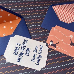 May include: Two dark blue envelopes with patterned liners, one featuring white flowers on orange and the other a red gingham check. A rectangular card with scalloped edges displays the text "HAVE A MEOW-VELOUS DAY! From, Your Crafty Friend" alongside a cat illustration.