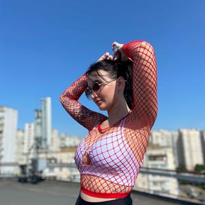 Puede incluir: Un top de red rojo sobre un sujetador deportivo blanco y leggings negros. El top tiene mangas largas y cuello redondo. El atuendo se usa al aire libre en un d&iacute;a soleado con un cielo azul claro de fondo.