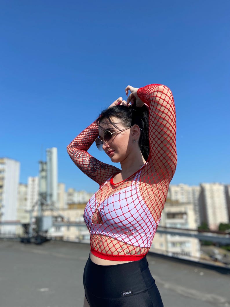 Puede incluir: Un top de red rojo sobre un sujetador deportivo blanco y leggings negros. El top tiene mangas largas y cuello redondo. El atuendo se usa al aire libre en un d&iacute;a soleado con un cielo azul claro de fondo.