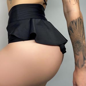 Könnte beinhalten: Eine schwarze High-Waist-Shorts mit einem breiten, glatten Bund und einem auffälligen, gerüschten Schößchen-Überrock, der sich um die Hüften legt. Das Kleidungsstück ist aus einem einfarbigen schwarzen Stoff mit leichtem Glanz gefertigt.