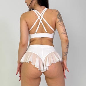May include: White lingerie set featuring a crisscross back bra and a ruffled skirted bottom. The bra has multiple straps and a classic clasp closure. The skirted bottom has a sheer, ruffled overlay, creating a layered effect. The set is made of a smooth, opaque fabric.