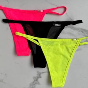 Pode incluir: Três cuecas de fio dental em cores vibrantes: rosa neon, preto e amarelo neon. A roupa íntima apresenta tiras finas e um design minimalista. A cueca preta tem um painel de malha transparente. Os itens são exibidos em uma superfície clara.