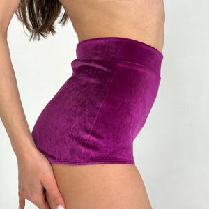 May include: A pair of vibrant purple velvet shorts. The shorts have a high waist and a soft, textured appearance. The fabric is a rich, saturated color, and the shorts appear to be well-fitted.