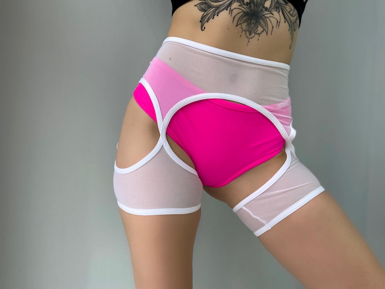 May include: A pair of athletic shorts featuring a bright pink bottom layer, overlaid with a sheer white mesh. White trim outlines the edges. The shorts have cut-out details on the sides.