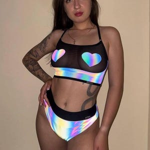May include: A two-piece set consisting of a black crop top and high-waisted bottoms. The top features a sheer black mesh upper section with two iridescent heart-shaped patches and an iridescent band. The bottoms have a matching iridescent finish.