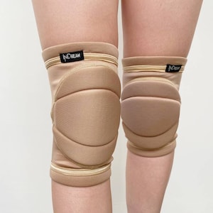 Può includere: Un paio di ginocchiere beige con un design imbottito. Hanno fasce elastiche beige con strisce dorate nella parte superiore e inferiore. Il marchio "INDREAM" è stampato su un'etichetta nera.