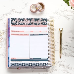 May include: A close-up of an open planner with a monthly budget page. The page is divided into sections for income, estimated expenses, actual expenses, and savings. The planner has a gold coil binding and a floral design on the cover.
