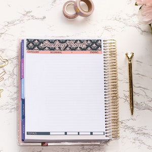 May include: A gold spiral bound planner with a black and pink floral design on the cover. The planner has a section labeled "Sinking Funds" and includes columns for category, beginning, ending, and totals. The planner is on a white marble surface with gold paper clips, a gold pen, and pink flowers.
