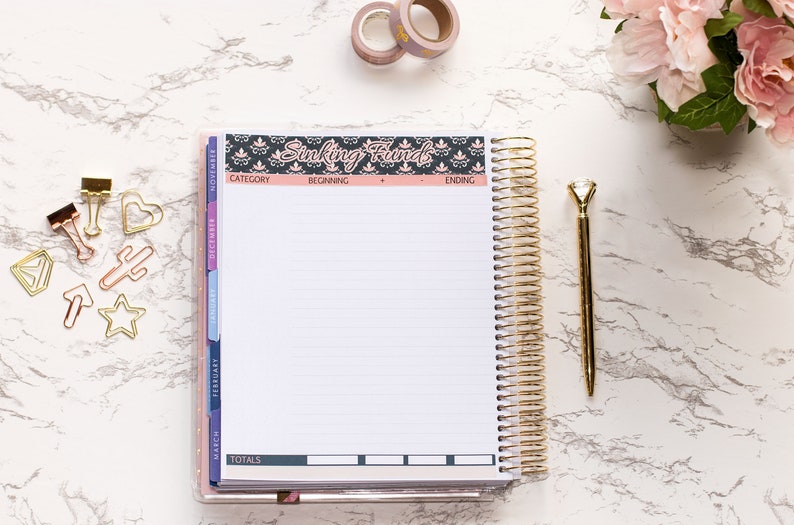 May include: A gold spiral bound planner with a black and pink floral design on the cover. The planner has a section labeled "Sinking Funds" and includes columns for category, beginning, ending, and totals. The planner is on a white marble surface with gold paper clips, a gold pen, and pink flowers.