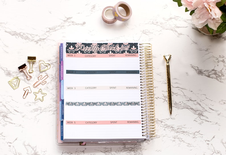 May include: A white planner with a gold spiral binding. The planner has a black and white patterned cover with the text "Weekly Check-ins". The planner is open to a page with the headings "Week 1", "Category", "Spent", and "Remaining".