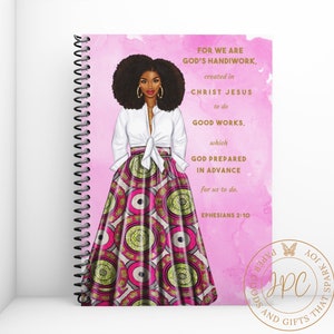 May include: A pink spiral bound notebook with a colorful illustration of a woman with an afro wearing a white shirt and a pink and green patterned skirt. The notebook has the text "FOR WE ARE GOD'S HANDIWORK, created in CHRIST JESUS to do GOOD WORKS, which GOD PREPARED IN ADVANCE for us to do. EPHESIANS 2-10" on the cover.