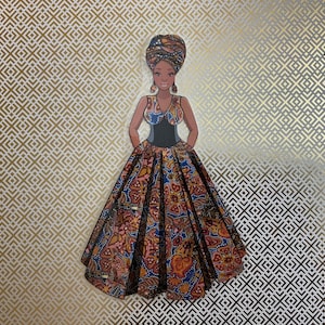 Op de afbeelding: Een papieren pop van een vrouw in een kleurrijke jurk en een hoofddoek. De pop staat op een gouden en witte gepatroneerde achtergrond.