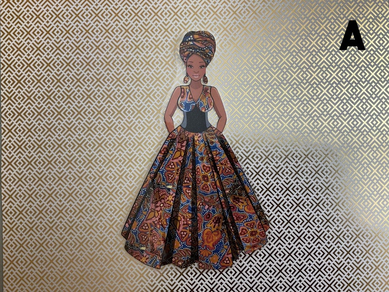 Op de afbeelding: Een papieren pop van een vrouw in een kleurrijke jurk en een hoofddoek. De pop staat op een gouden en witte gepatroneerde achtergrond.