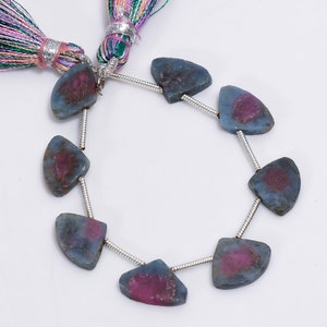 May include: Strand of gemstone beads. Each bead is a unique, semi-circular shape with a blue-gray exterior and a vibrant magenta center. The beads are strung on a silver-colored wire. Colorful tassels are visible in the background.