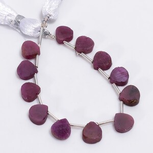 Op de afbeelding: Een ketting van hartvormige, diep magenta edelstenen. De edelstenen zijn geregen op een zilverkleurige draad, met een witte kwast aan het uiteinde. De edelstenen hebben een natuurlijk, getextureerd uiterlijk.