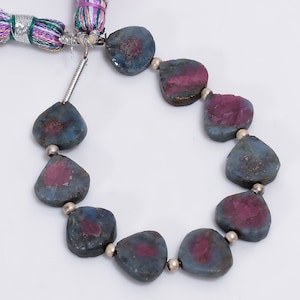 May include: A necklace of heart-shaped, blue-grey gemstones with magenta centers. The beads are strung with small silver beads. A colourful tassel is attached to the necklace.