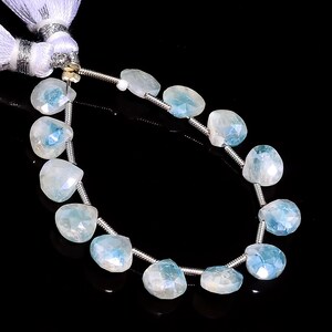 May include: A strand of faceted, heart-shaped aquamarine beads. The beads are a pale blue color with internal inclusions, strung on a silver-colored wire. White tassels are attached to the end of the strand. The background is black.