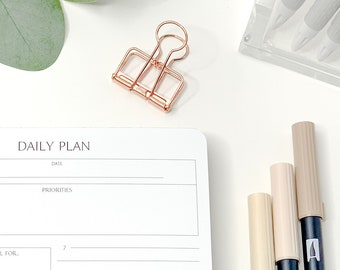 May include: A white daily planner with the text "DAILY PLAN" and sections for priorities and tasks. A clear pen, Tombow ABT markers, and a rose gold binder clip are also visible. The planner is from Yesenia Diaries.