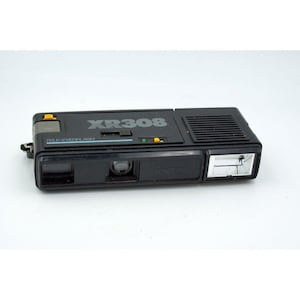 May include: Black Keystone XR308 Tele-Everflash camera. The camera has a flash, a lens, and the text "XR308" and "Keystone" printed on it. The camera has a yellow button and a black strap attachment.