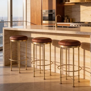 May include: Three bar stools with circular brown leather seats and brushed gold metal frames featuring four vertical legs and a circular footrest, positioned in front of a kitchen island with a light-colored stone countertop and base.