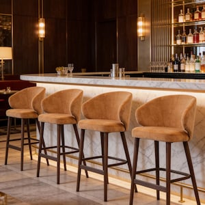 Pode incluir: Quatro bancos de bar com assentos curvos estofados em tom bronzeado e pernas de madeira castanho-escura. Cada banco apresenta um encosto arredondado e um apoio para os pés, alinhados ao longo de um balcão de bar em mármore branco com luz.