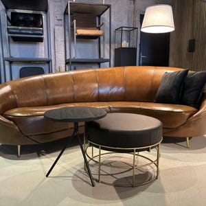Può includere: Un divano in pelle marrone dal design curvo, abbinato a un tavolino nero e un pouf rotondo. Due cuscini neri poggiano sul divano. L'arredamento ha un'estetica moderna con gambe color oro.