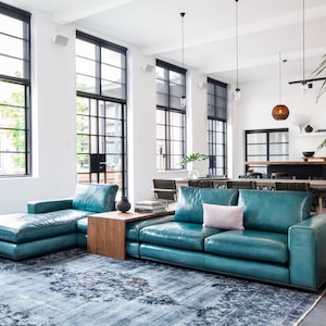 Könnte beinhalten: Modernes Wohnzimmer mit einem teal-farbenen Ledersofa und einer Chaiselongue. Der Raum verfügt über große Fenster mit schwarzen Rahmen, einen gemusterten Teppich und Beistelltische aus Holz. Dekorative Hängelampen und Pflanzen ergänzen das moderne Design.