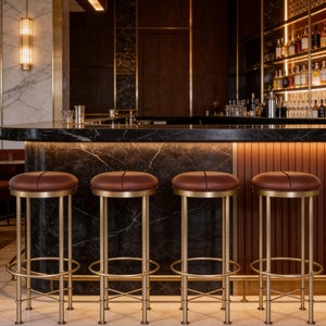 May include: Four bar stools with round, dark brown leather-upholstered seats and brushed gold metal frames. Each stool features a circular footrest and four slender legs connected by a cross-brace structure, positioned in front of a marble bar.